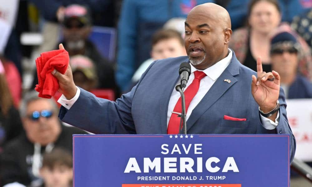 North Carolina lieutenant governor Mark Robinson wears a white button up shirt, red tie, blue suit jacket and red pocket square as he stands at a podium with a sign reading 'Save America' on it. He is gesturing with both his hands in the air and one hand is holding a red towel