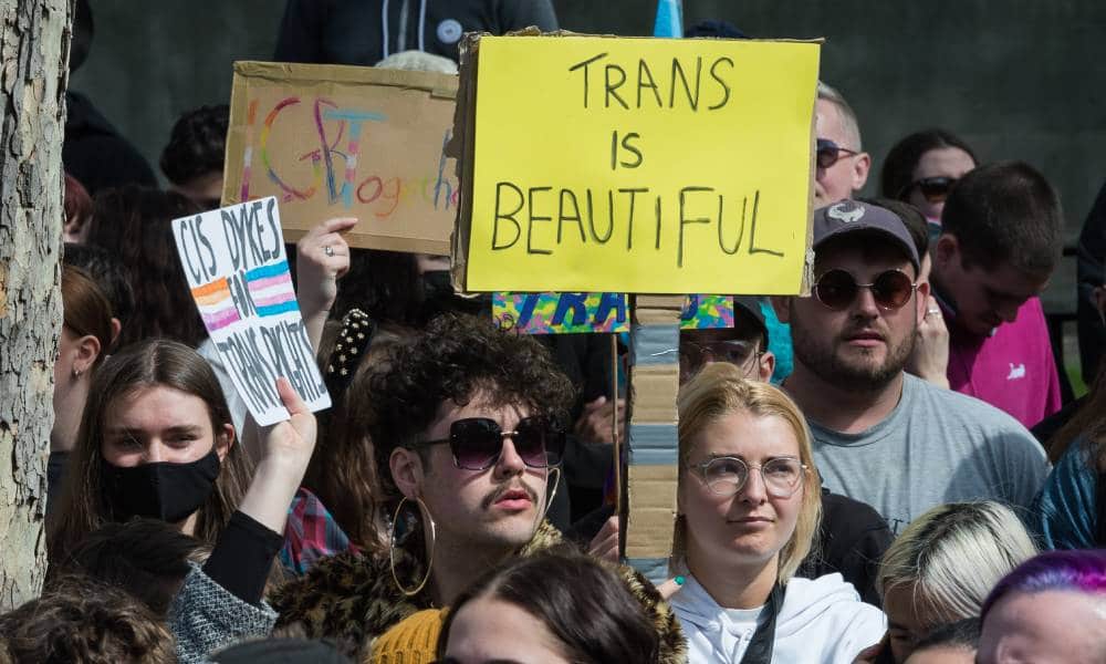 Several people gather in a crowd in support of the trans community in the UK. One person holds up a sign reading 'trans is beautiful' in the crowd.
