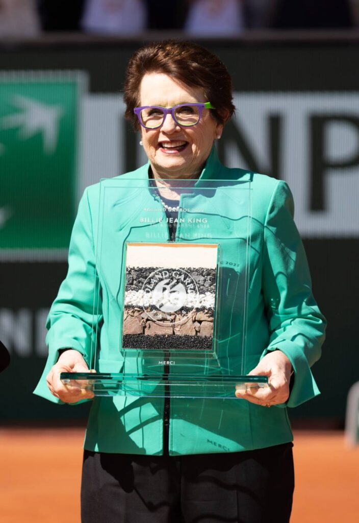Billie Jean King stands on the court of the French Open tennis championship while wearing a bright green jacket and dark bottoms. She is holding an award which contains multiple layers of sediment in a clear square container that has her name written on it in white