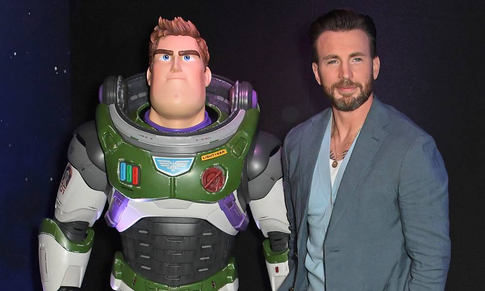 Chris Evans wears white shirt, light blue sweater and grey-blue suit jacket with matching trousers as he stands next to a statue of the Disney character Buzz Lightyear. Buzz is a character that wears a white, green and purple space suit with a panel on the chest