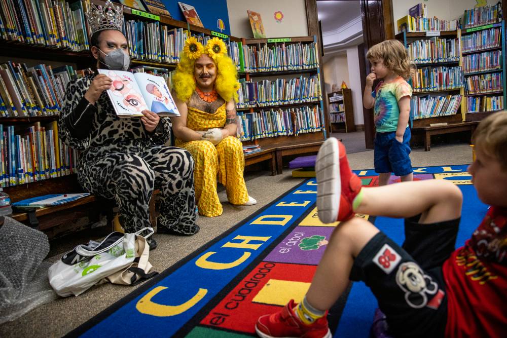 Two drag performers read from a children's book in front of several young children during an event at a library