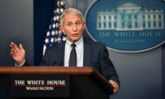 Anthony Fauci stands at a podium in the White House with the White House logo and an American flag in the background. Fauci is wearing a white shirt, dark tie and dark suit jacket as he talks to people off camera