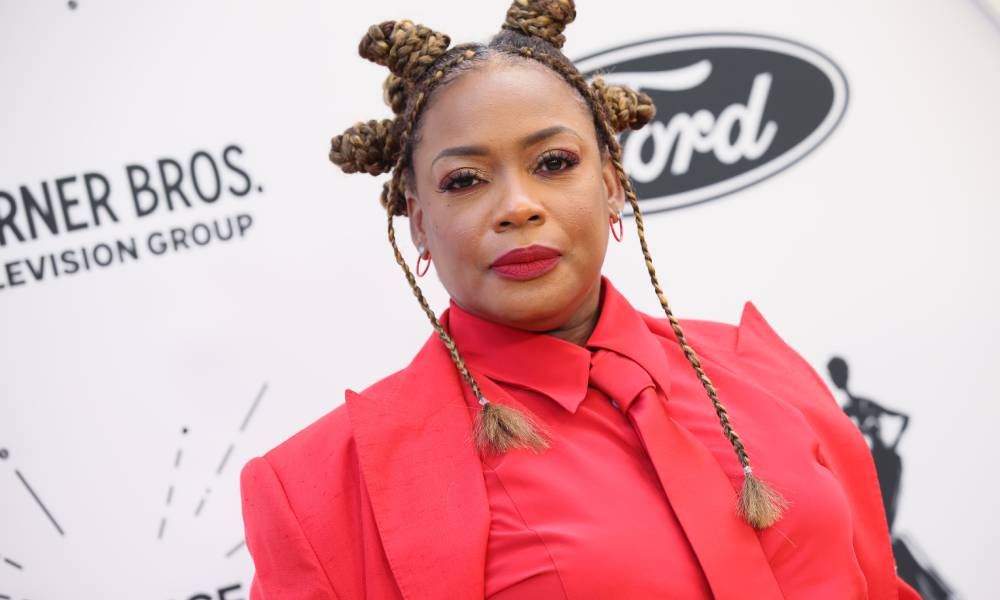 Aunjanue Ellis wears a bright red suit jacket, matching red button up shirt and tie. She also wears matching red lipstick and has her braided hair up in knotted designs on her head