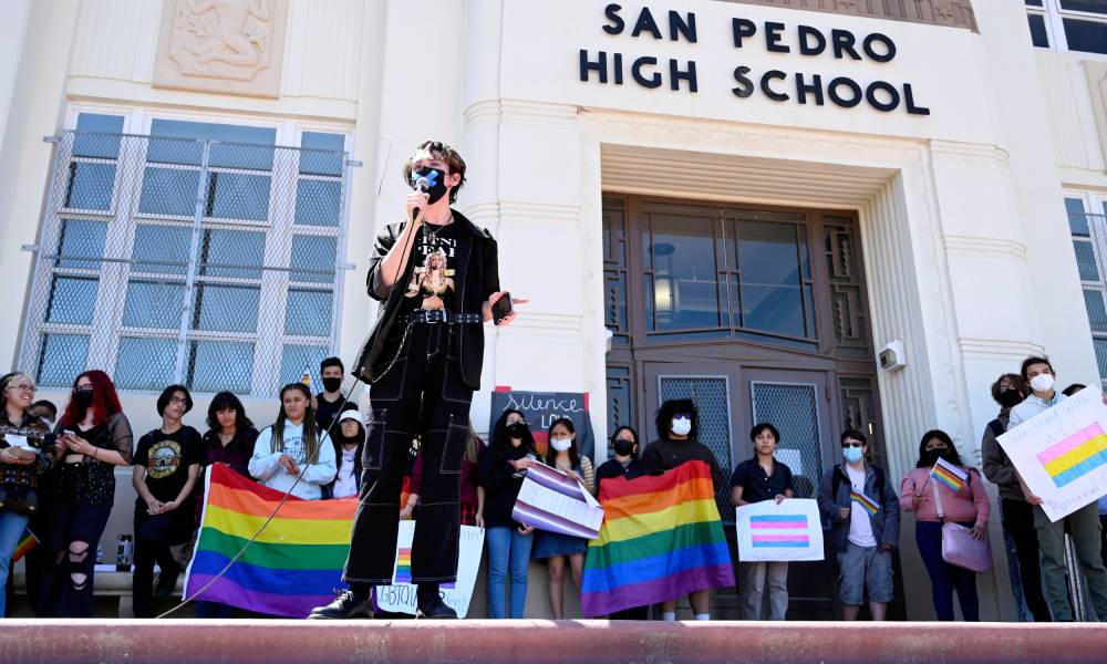 One high school student who is wearing a face mask speaks into a microphone as other young people stand behind them all holding LGBTQ+ flags, signs supporting the LGBTQ+ community. The group is standing on the steps to San Pedro High School