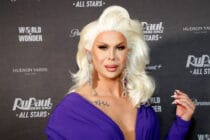 Semi-close up photo of Trinity the Tuck attending the 'Drag Race All Stars' premiere. They are wearing a platinum blonde wig, and a dark purple dress.