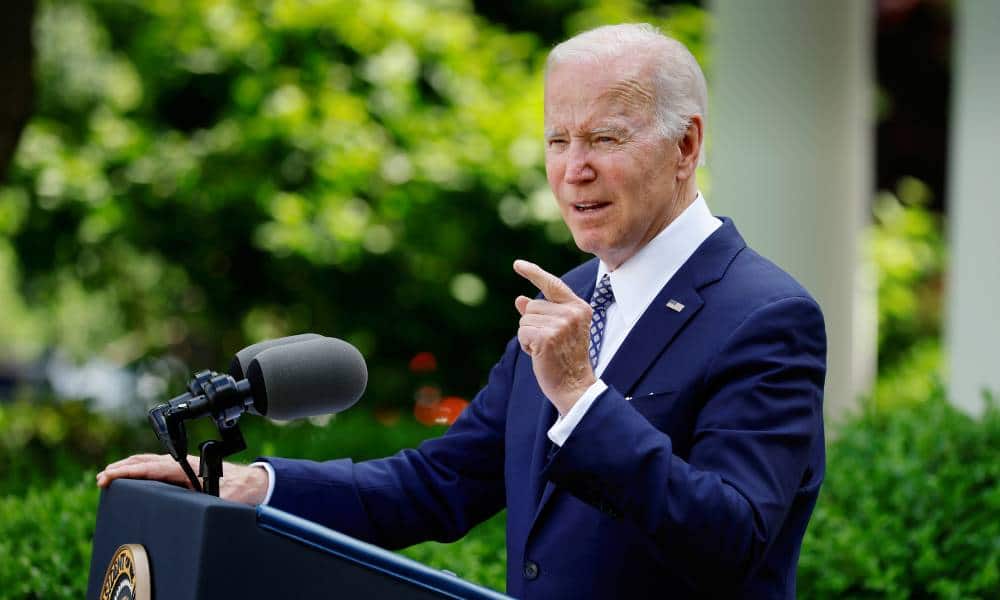 President Joe Biden stands at a podium while outside while wearing a white button up shirt, blue suit jacket and tie. He is pointing upwards with one finger on one hand