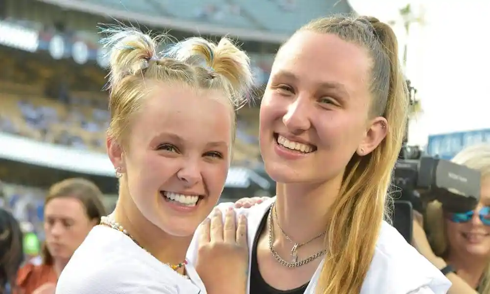 JoJo Siwa smiles at the camera while wearing a white Dodgers jersey. She has her hands on the shoulder of Kylie Prew who is also wearing a white Dodgers jersey, ponytail and silver necklaces 