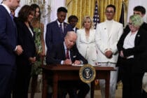 President Biden sits at a desk, signing an executive order, while LGBT+ advocates watch