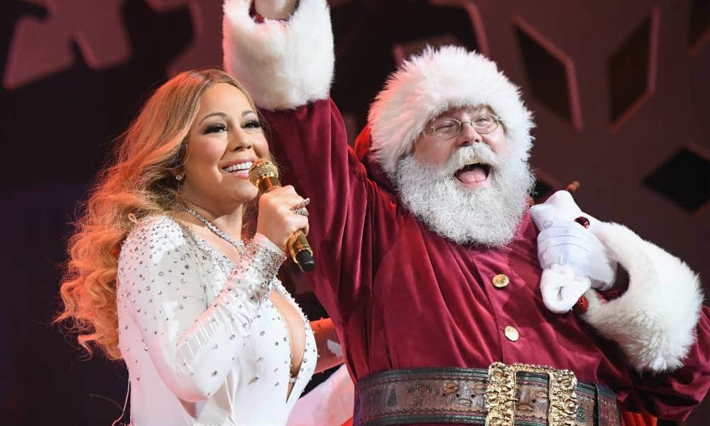 Mariah Carey sings into a golden microphone while wearing a white shirt with rhinestones on it. She is accompanied on stage by a person dressed as Santa Claus in a red and white suit with a big brown belt and matching red and white hat