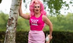 Drag Race UK star Ella Vaday wears a pink wig, hot pink sleeveless shirt with a white 'Race for Life' logo on it and baby pink short skirt. She poses with one hand on a tree while holding one foot, which has a white high heel on it, in her other hand