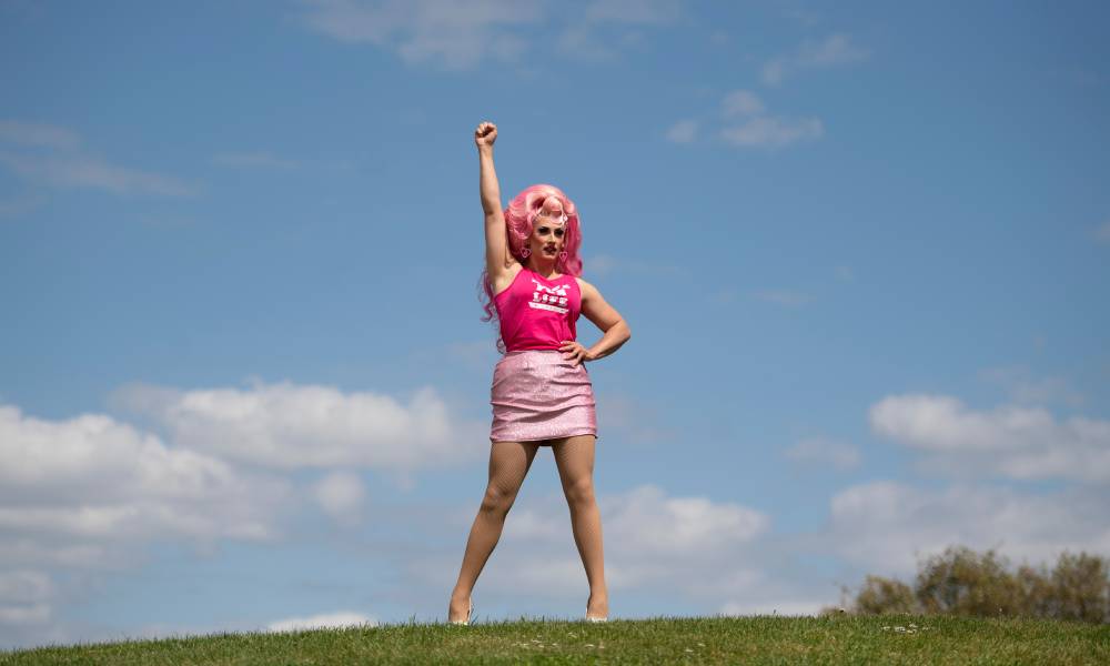 Drag Race UK star Ella Vaday wears a pink wig, hot pink sleeveless shirt with a white 'Race for Life' logo on it and baby pink short skirt. She poses on top of what appears to be a hill with one hand raised up in the air while her other hand rests on her hip