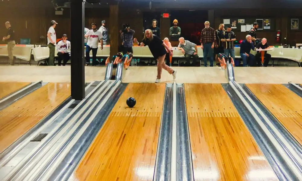 Michelle Guzowski wears a black shirt and tan trousers as she throws a dark bowling bowl down a wooden lane in a bowling alley