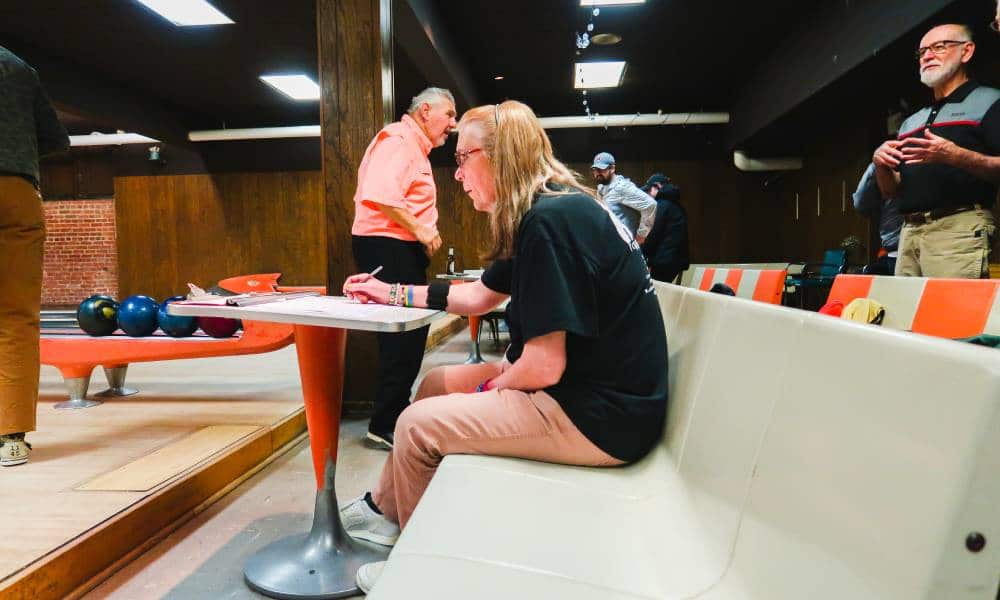 Michelle Guzowski sits on a white bench in a bowling alley and is writing on a piece of paper before her. She is wearing a black top and tan bottoms