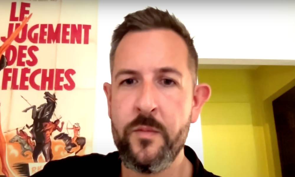 James McFadzean sits in a room wearing a dark shirt. The wall behind him is white and there is a poster with a person rising a bucking horse with words in French written on it