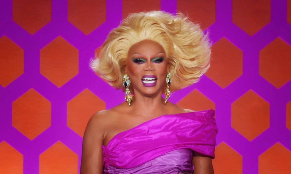RuPaul laughs while wearing a pink off the shoulder dress, blonde wig and silver sparkly earrings while on the set of Drag Race All Stars 7