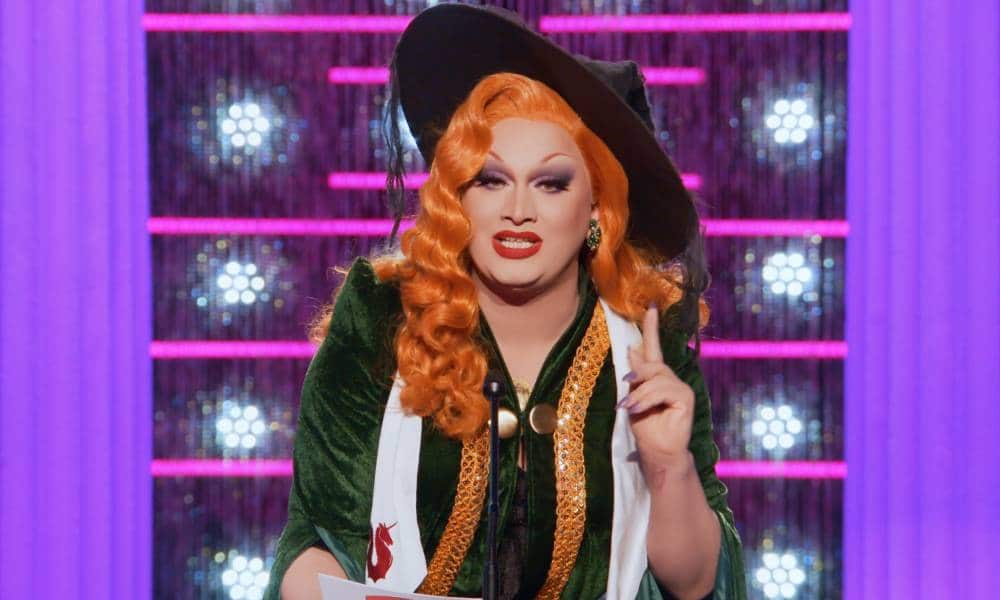 Drag Race All Stars 7 star Jinkx Monsoon wears a long orange wig, black witch's hat, green velvet graduation gown and white fabric over her neck as she stands at a podium and gestures with one finger