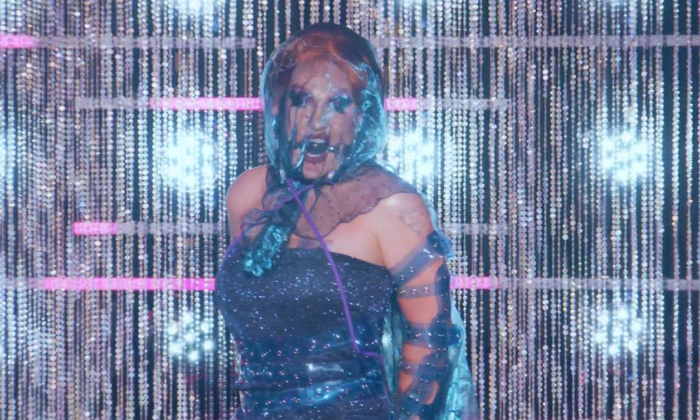 Drag Race All Stars 7 star The Vivienne wears a blue mesh veil and matching blue dress with transparent buckles all over her body with a ball-gag in her mouth