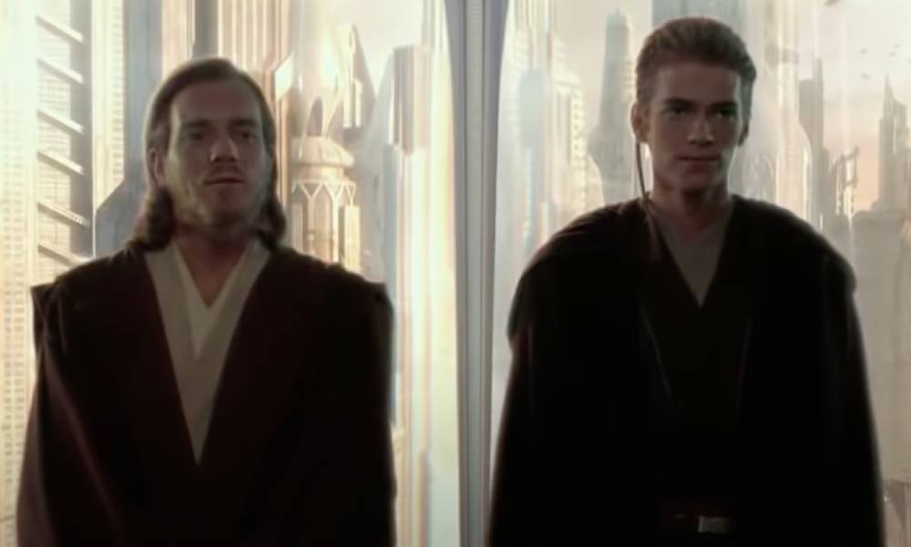 Ewan McGregor plays Obi-Wan Kenobi who is wearing a dark robe and tan clothing underneath. He is standing beside Anakin Skywalker, played by Hayden Christensen, who is also wearing dark coloured robes with a futuristic cityscape seen in the background