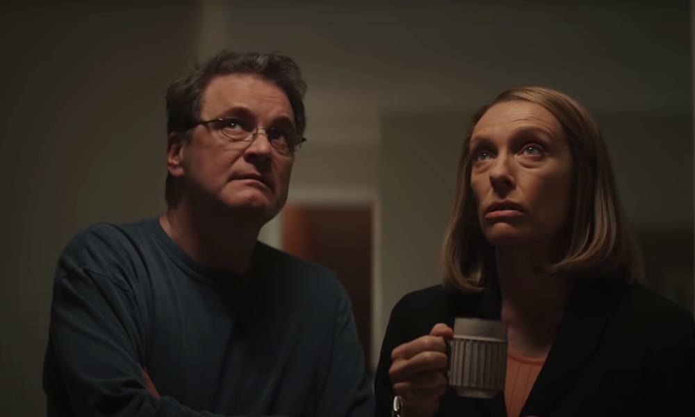 Colin Firth wears a dark top and glasses as he plays Michael Peterson in HBO Max's series The Staircase. Toni Collette stands next to him wearing a dark top as well and holding a cup as she portrays Peterson's wife Kathleen. Both actors are looking up at something