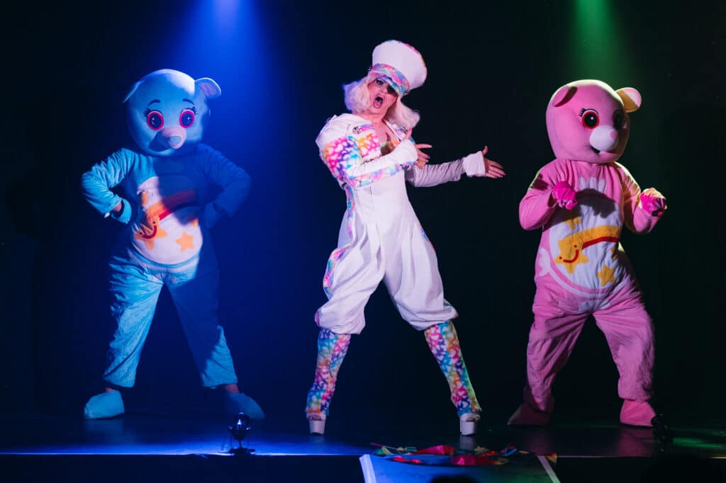 A drag performer flanked by two performers in blue and pink teddy bear costumes