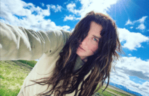 A selfie of Kesha. She's holding the camera, wearing a light hoody, standing in front of a blue but cloudy sky