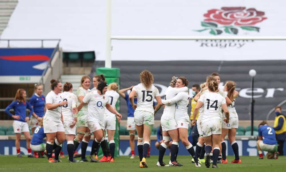 Several women rugby players wearing white gather around in celebration after they win a match. Some of hugging each other while others are running towards the crowd