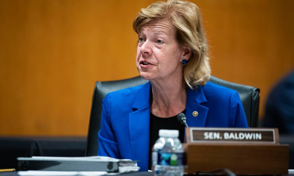 Senator Tammy Baldwin sits down behind a desk while wearing a black top and blue blazer jacket with a small golden pin on it