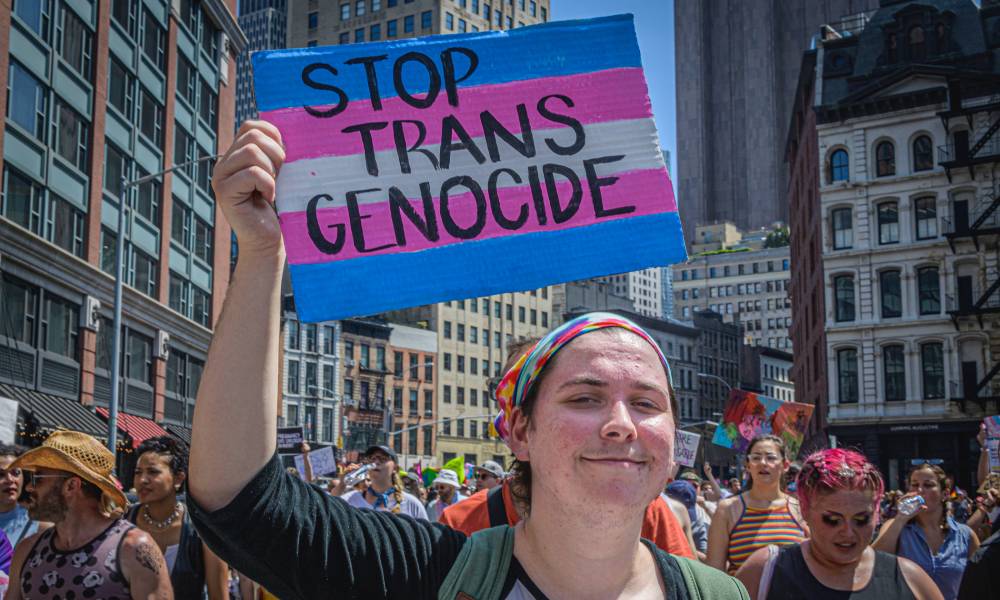 Participant seen holding a sign in the colours of the trans Pride flag that reads "Stop trans genocide" at the protest.