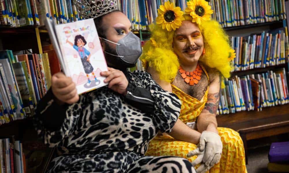 One drag performer wears a monochromatic animal print outfit, sparkly face mask and silver crown as they read from a children's book held up in their hands. Another queen dressed in a bright yellow patterned outfit with a matching yellow wig with two sunflowers sits beside the other performer