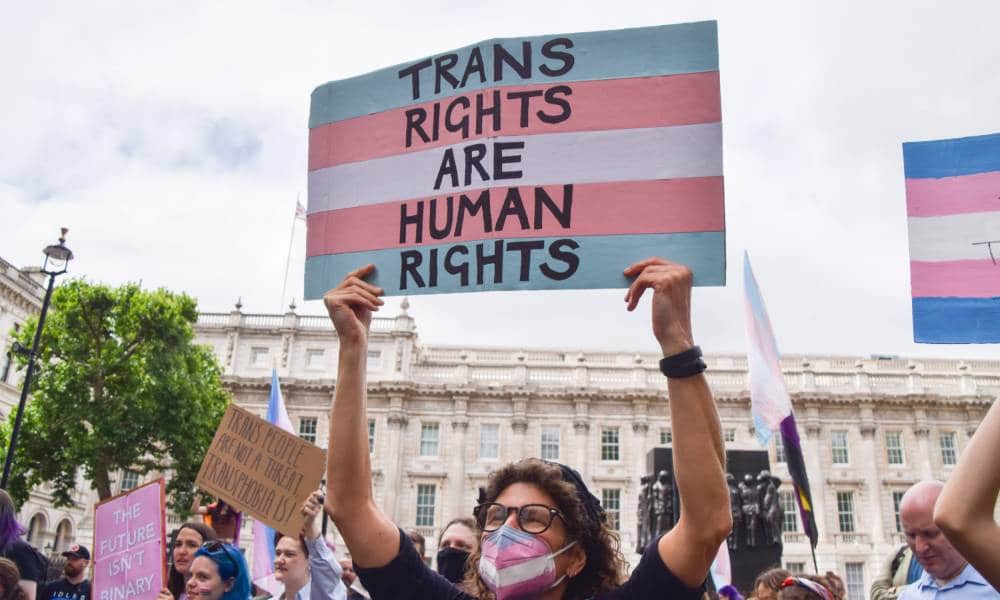 A person wearing a face mask designed in the colours of the trans Pride flag holds up a sign with the colours of the trans Pride flag that reads "Trans rights are human rights" above their head during a protest