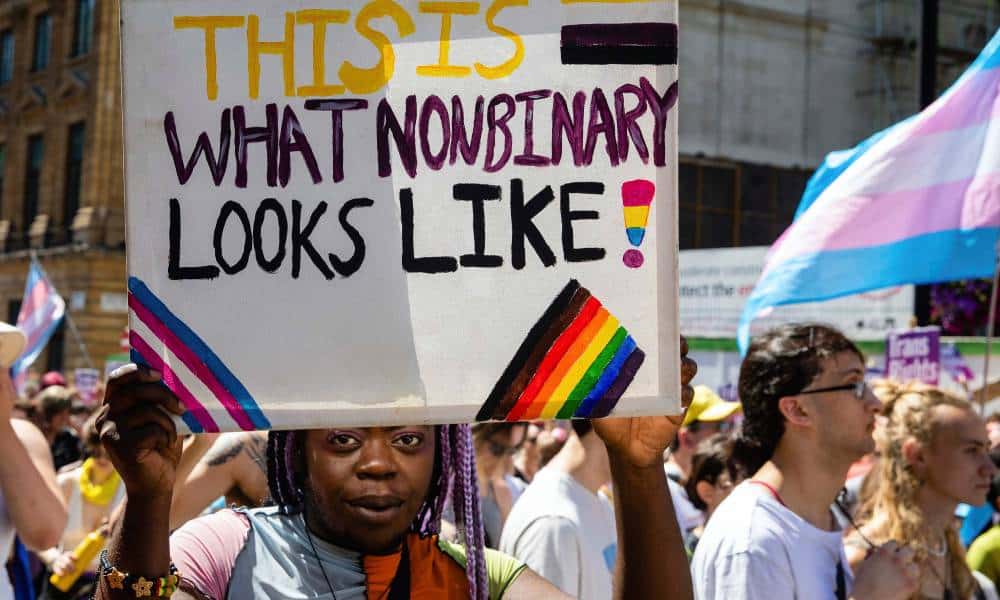 A person holds up a white sign with the words 'This is what nonbinary looks like' written in yellow, purple and black colours