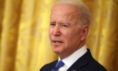 President Joe Biden is seen wearing a white button up shirt, blue tie and dark suit jacket as he stands in front of a yellow background