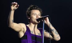 Harry Styles wears a black and purple outfit as he speaks into a microphone and holds one arm in the air