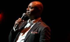 Dave Chappelle wears a white button up shirt, dark suit jacket and has something sticking out of the jacket's breast pocket as he speaks into a microphone he is holding is his hand