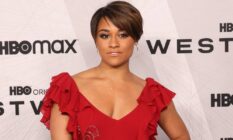Ariana DeBose wears a red dress with frills on the sleeves as she stands in front of a white background with black logos for HBO Max and Westworld