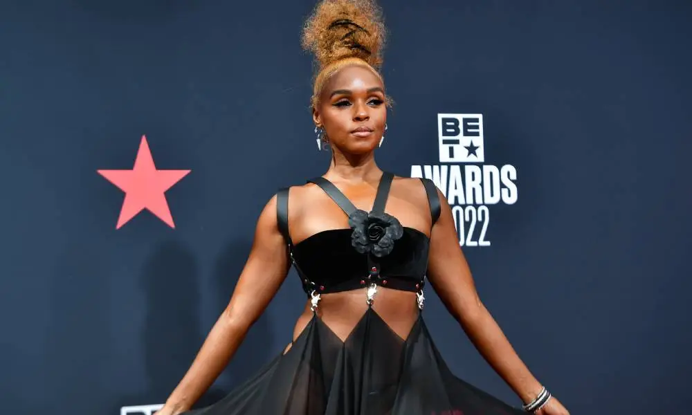 Janelle Monáe posts while wearing a black dress consisting of straps and drapped fabric with their hair styled on top of their head