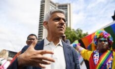 Major of London Sadiq Khan wears a white button up shirt and grey jacket as he attends Pride in London celebrations. A person in the background wears rainbow clothing as they hold up an LGBTQ+ Pride flag
