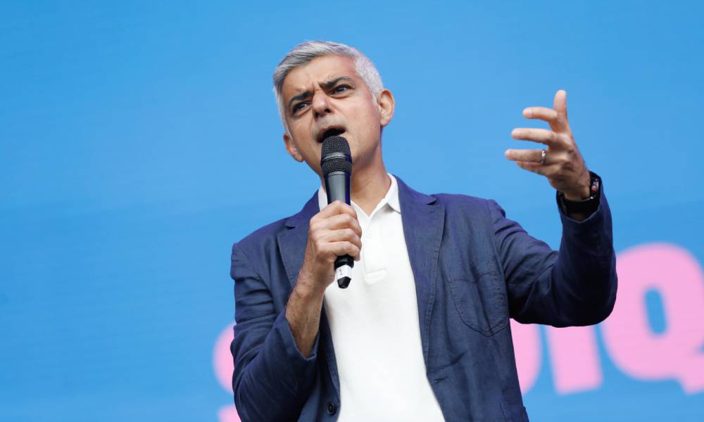 Mayor of London Sadiq Khan wears a white button up shirt and blue jacket as he holds a microphone that he is speaking into and stands in front of a blue background with pink lettering