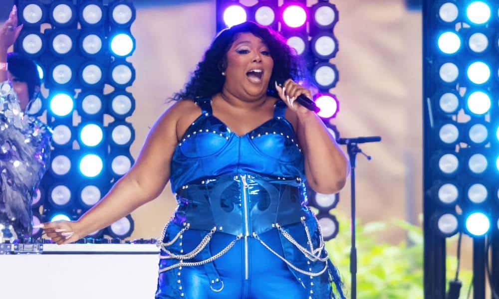 Lizzo wears a blue reflective outfit with silver chains hanging from her hips as she sings into a microphone as she performs on stage