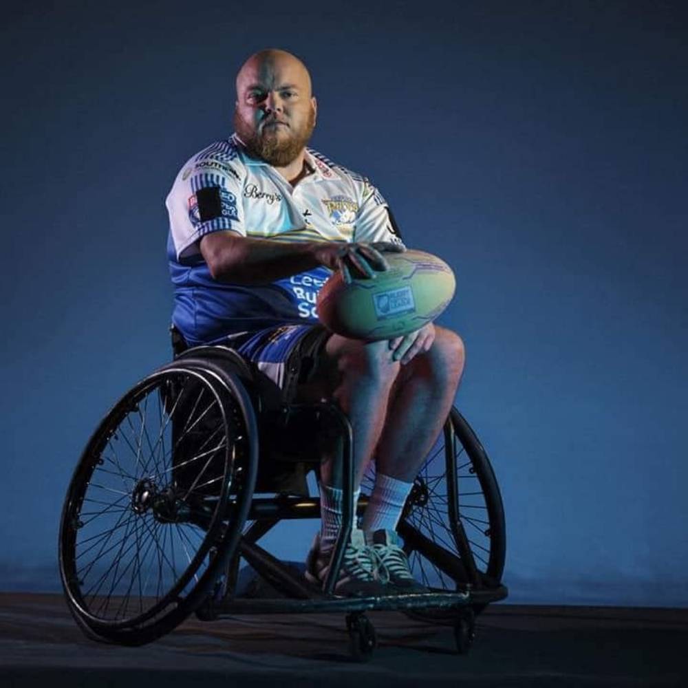 Verity Smith, un joueur de rugby, porte un uniforme blanc alors qu'il est assis dans un fauteuil roulant tout en tenant un ballon de rugby sur ses genoux