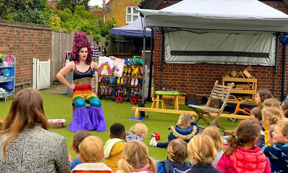 Aida H Dee, a drag queen and founder of Drag Queen Story Hour UK, wears a black top and rainbow bottoms as she reads from a book in front of a crowd of children gathered outside