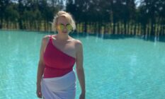 Rebel Wilson wears a pink off the shoulder swimsuit with a white cloth around her hips as she stands near a pool