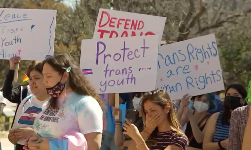 A crowd of people gather in support of the trans community in Texas. One person holds up a sign reading 'Protect trans youth' while another has a sign reading 'Trans rights are human rights'
