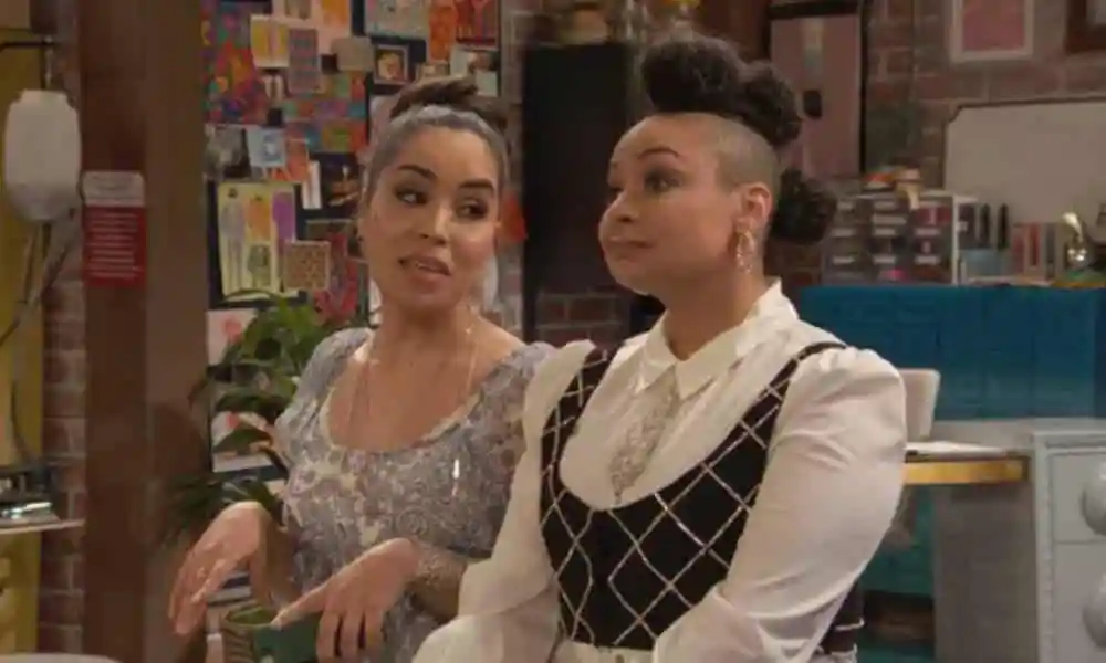 Raven's Home character Nikki (Juliana Joel) wears a white dress with a blue damask pattern on it as she speaks to Raven Baxter (Raven-Symoné). Raven is wearing a white button up shirt, glittery small tie and black and silver patterned vest. Her hair is styled up in three bunches running down her head.