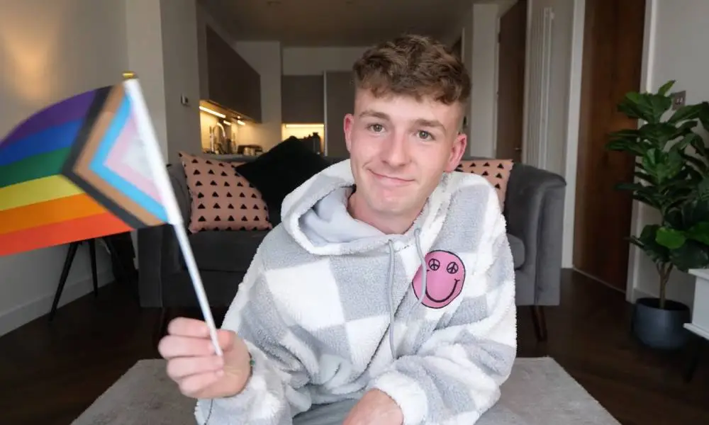 Adam Beales, known as Adam B on YouTube, wears a grey and white checkered fuzzy sweatshirt with a pink smiley face on the chest. He is waving a progressive Pride flag in one hand.