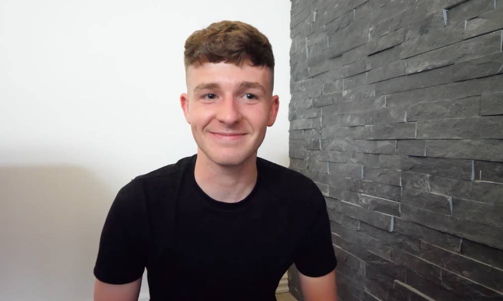 Adam Beales, known as Adam B on YouTube, sits next to a grey brick wall as he wears a black t-shirt