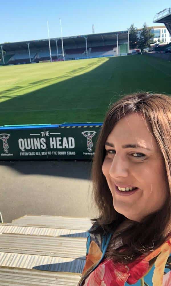 Emily Hamilton porte une tenue à motifs floraux alors qu'elle se tient devant un terrain de rugby avec les mots 'Quin's head' visibles derrière elle