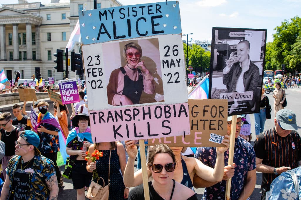 Alice Litman's family marching in a thick crowd, holding two placards. The first is in the colour of the trans flag, has a photo of Alice and the words: 'My sister Alice, transphobia kills, 23 Feb '02 - 26 May '22'. The second has a black and white photo Alice, and says 'In loving memory of Alice Litman'. 