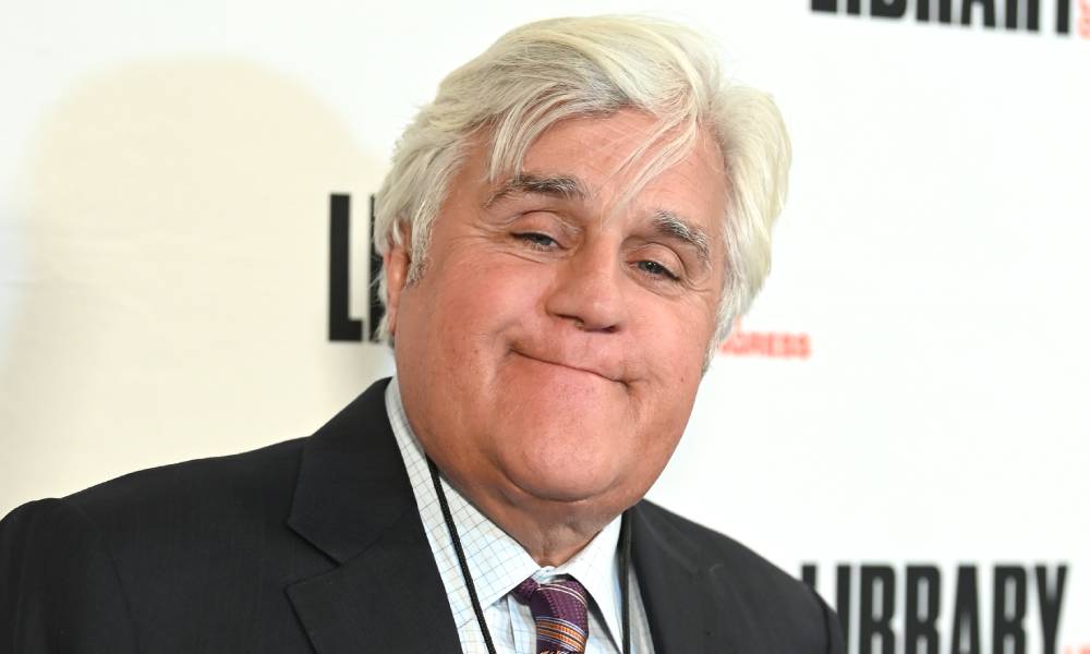 Jay Leno smiles at the camera as he wears a white shirt, patterned tie and black suit jacket