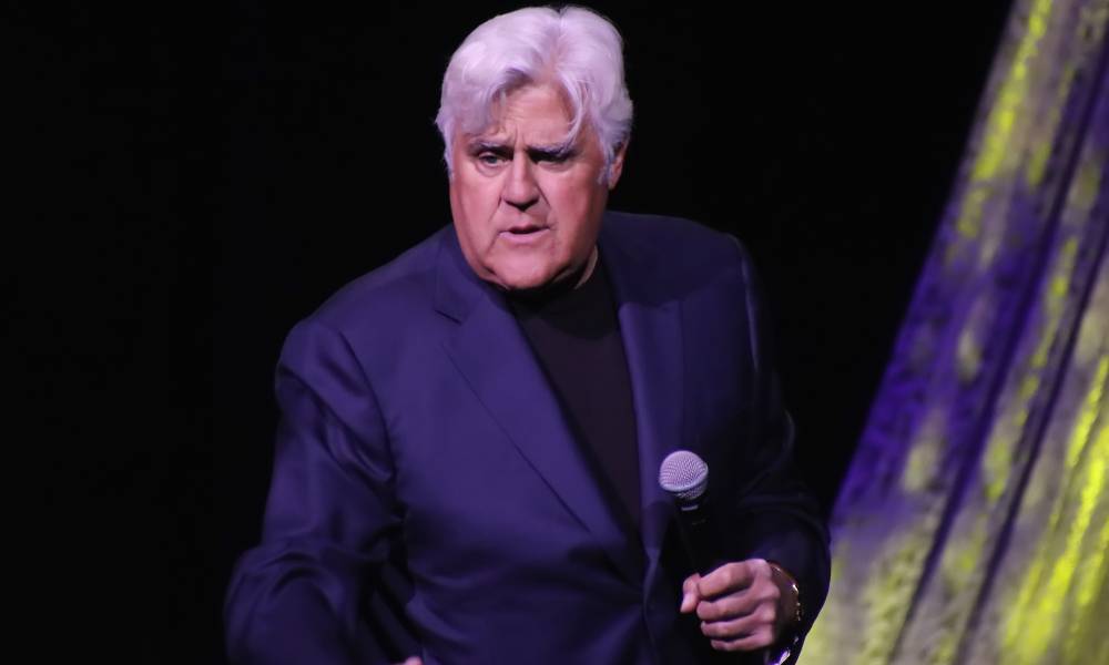 Jay Leno holds a microphone in his hand as he wears a black shirt and dark blue suit jacket as he stands on stage